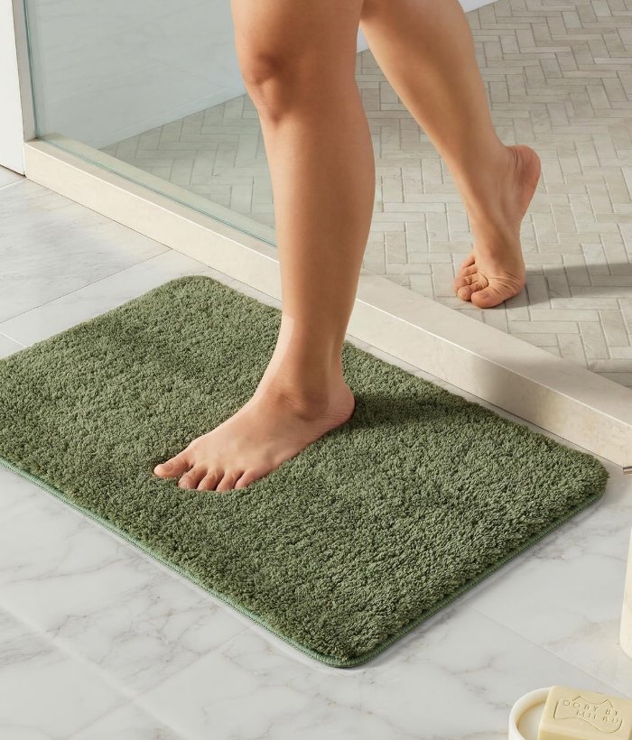 A sage TYUU bath mat on warm tile with bare feet stepping onto it